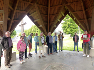 Eine Gruppe unterschiedlicher Erwachsener steht unter einem Holzpavillon, einige betrachten eine Bodenplatte, andere die Umgebung.