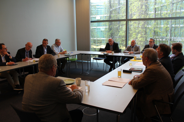 Männer in Anzügen bei einem Meeting an einem langen Tisch in einem hellen Konferenzraum mit großen Fenstern.