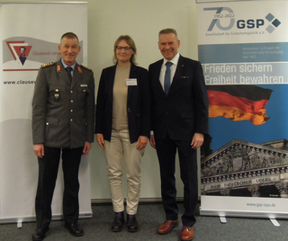 Ein Soldat in Uniform, eine Frau und ein Mann im Anzug lächeln vor Bannern der Clausewitz-Gesellschaft und GSP.