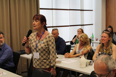 Eine Frau spricht mit einem Mikrofon bei einer Konferenz, während andere Teilnehmer an Tischen im Hintergrund sitzen.