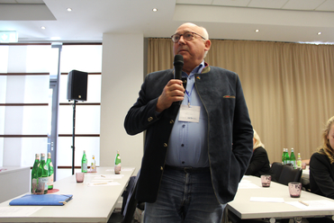 Ein Mann mit Brille und Trachtenjacke spricht in ein Mikrofon bei einer Präsentation oder Konferenz.