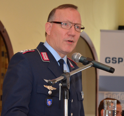 Ein deutscher Militäroffizier in Uniform und Brille spricht in ein Mikrofon. Pilotenschwingen und Hessenwappen sind zu sehen.
