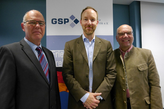 Drei lächelnde Männer, einer in Trachtenjacke, posieren vor einem Banner der Gesellschaft für Sicherheitspolitik (GSP).