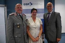 Drei lächelnde Personen: ein Mann in Militäruniform, eine Frau im hellen Kleid und ein Mann im Anzug, vor einem GSP-Banner.