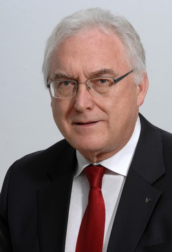 Porträt eines älteren Mannes mit weißem Haar, Brille, dunklem Anzug und roter Krawatte, der direkt in die Kamera blickt.