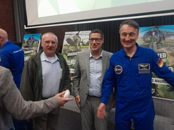 Drei lächelnde Männer, darunter ein ESA-Astronaut im blauen Overall, posieren vor Postern mit Hubschraubern.