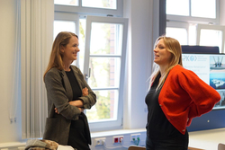 Zwei lächelnde Frauen in einem Innenraum, eine im Karo-Blazer, die andere im roten Cardigan, im Gespräch vertieft.