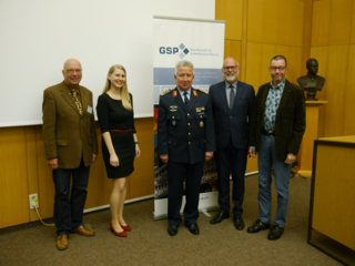 Fünf Personen, darunter ein Soldat in Uniform, posieren lächelnd vor einem Banner der Gesellschaft für Sicherheitspolitik e.V. bei einer Veranstaltung.