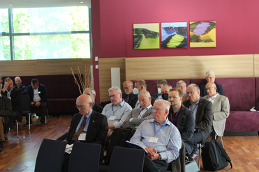 Eine Gruppe hauptsächlich älterer Männer nimmt an einer Konferenz oder einem Seminar in einem Raum mit großen Fenstern und Holzboden teil.