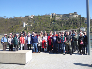 Große Gruppe von Personen posiert für ein Foto auf einer Promenade mit historischer Festung und Seilbahnen im Hintergrund.