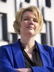 Porträt einer blonden Frau im blauen Blazer mit verschränkten Armen, die selbstbewusst in die Kamera blickt, unscharfes Gebäude im Hintergrund.