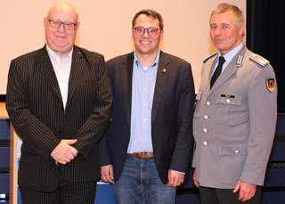 Drei Männer stehend: einer im Nadelstreifenanzug, einer in Blazer und Jeans, und einer in grauer Militäruniform.