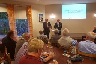 Zwei Redner präsentieren einer Gruppe von Männern in einem Raum, mit 'Herzlichen Dank für die Aufmerksamkeit!' auf dem Beamer.