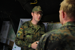 Soldat Dr. Porsch in deutscher Tarnuniform mit NATO-Abzeichen im Gespräch mit einem Kollegen in einem Zelt.