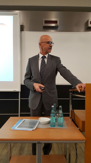 Ein Mann im Anzug mit Brille und Schnurrbart präsentiert in einem Raum mit Bildschirm und kWh-Anzeigen.