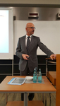 Ein Mann im Anzug mit Brille und Schnurrbart präsentiert in einem Raum mit Bildschirm und kWh-Anzeigen.