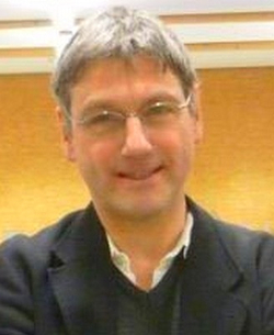 Porträt eines lächelnden Mannes mittleren Alters mit grauem Haar und Brille, der eine dunkle Jacke trägt.