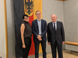 Drei lächelnde Personen, eine Frau, ein junger und ein älterer Mann, stehen vor einer Deutschlandflagge mit Bundesadler.