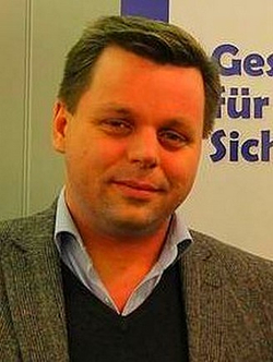 Nahaufnahme eines Mannes mittleren Alters mit kurzen braunen Haaren, Tweedjacke, blauem Hemd und dunklem Pullover, leicht lächelnd.