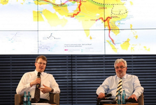 Zwei Männer diskutieren vor einer Karte der chinesischen Belt-and-Road-Initiative, die Handelsrouten und Pipelines zeigt.