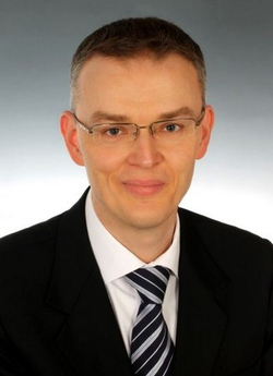 Porträt eines hellhäutigen Mannes mit kurzen Haaren und Brille, der einen dunklen Anzug und eine gestreifte Krawatte trägt, leicht lächelnd.