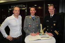 Drei lächelnde Männer, zwei in Uniform (Heer, Marine) und einer in Zivil, posieren vor einer Bibliothek. Einer unterschreibt ein Gästebuch.