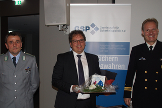 Drei lächelnde Männer, zwei in Uniformen und einer im Anzug mit einem Geschenk, stehen vor einem GSP-Banner.