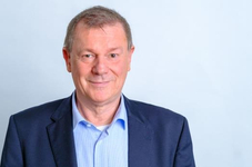 Ein lächelnder mittelalter Mann in blauem Hemd und dunklem Blazer schaut direkt in die Kamera. Heller Hintergrund.
