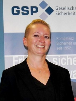 Lächelnde Frau mit blonden Haaren und schwarzem Blazer, vor einem GSP-Logo-Banner stehend.