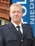 Ein lächelnder Mann mit rötlichem Haar in dunkler Polizeiuniform, weißem Hemd und Krawatte, vor einem "Polizei Niedersachsen"-Schild.
