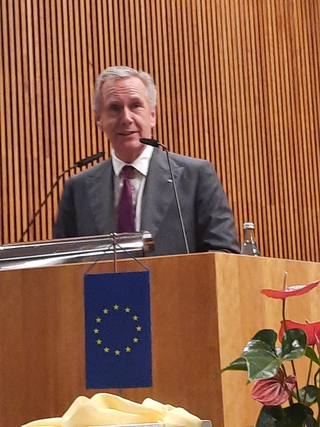 Ein grauhaariger Mann im Anzug hält eine Rede an einem Holzpult mit zwei Mikrofonen, einem EU-Banner und einer Pflanze.