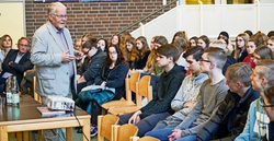 Ein älterer Mann spricht zu einer sitzenden Gruppe junger Menschen, wahrscheinlich Schüler, in einem Innenraum.