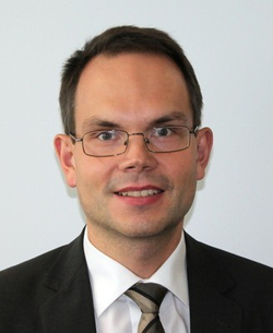 Ein lächelnder Mann mit kurzen dunklen Haaren und Brille, trägt einen dunklen Anzug und gestreifte Krawatte in einem professionellen Porträt.