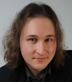Kopfbild eines Mannes mit langen braunen Haaren, dunkler Jacke, blickt in die Kamera.