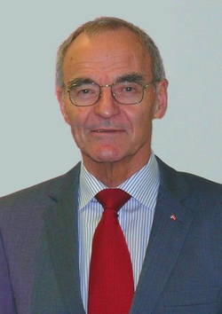 Porträt eines älteren Mannes mit Brille, grauem Anzug, gestreiftem Hemd, roter Krawatte und kleiner rot-weiß-roter Anstecknadel.