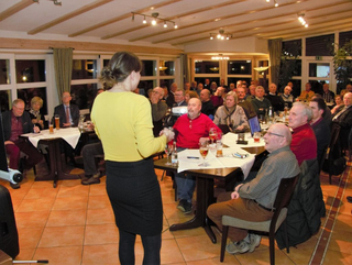 Eine Frau spricht vor einem Publikum älterer Erwachsener an Tischen bei einer Veranstaltung oder Präsentation.
