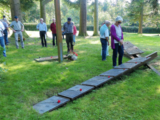 Gruppe älterer Menschen an Gedenktafeln mit roten Blumen auf einem Rasenfeld unter Bäumen bei einer Gedenkfeier.
