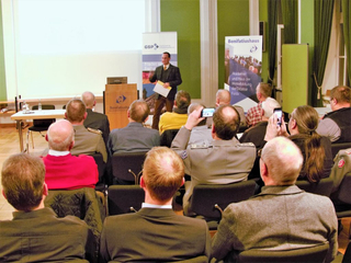 Ein Mann hält einen Vortrag vor einem Publikum, einige filmen mit Handys. Bonifatiushaus-Banner sichtbar.