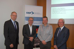 Vier lächelnde Männer posieren vor einem GSP-Banner und einer Projektionsfläche bei einer sicherheitspolitischen Veranstaltung.