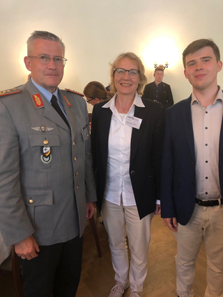 Ein Militäroffizier, eine Frau mit Brille und GSP-Namensschild sowie ein junger Mann im Blazer posieren drinnen für ein Foto.