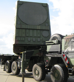 Ein großer, militärischer Mehrrad-LKW transportiert ein massives, olivgrünes Radarsystem mit einer runden, hexagonal gemusterten Antenne.