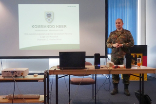 Ein deutscher Heeresoffizier in Tarnuniform präsentiert zur Ausrüstungsplanung. 'KOMMANDO HEER' wird auf der Leinwand gezeigt.