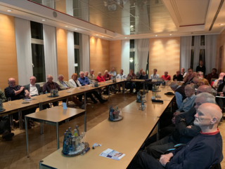 Ein Raum voller Menschen, hauptsächlich ältere Erwachsene, die an langen Tischen sitzen und eine Versammlung oder Präsentation besuchen.