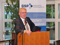 Mann im Anzug spricht an einem Holzpult mit Mikrofon, dahinter ein GSP-Banner für Sicherheitspolitik.