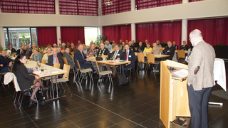Zuhörer bei einer Veranstaltung in einem großen Raum mit roten Vorhängen; ein Redner steht rechts an einem Pult.