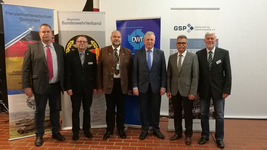 Sechs Männer in Anzügen und Jacken posieren vor Bannern von Militär- und sicherheitspolitischen Organisationen.