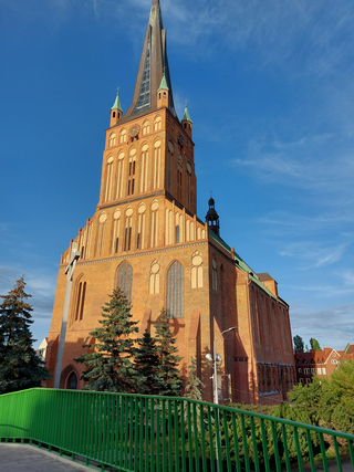 Backsteinkirche mit hohem, spitzem Turm, grünen Bäumen und einem grünen Geländer im Vordergrund, unter blauem Himmel bei Sonnenschein.