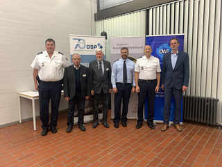 Sechs Männer, teils in Uniform, teils in Anzügen, posieren vor Bannern von Sicherheits- und Militärverbänden.