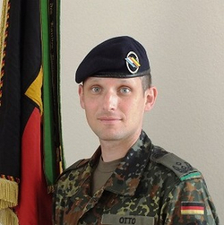 Porträt des deutschen Soldaten Otto in Flecktarn-Uniform, dunkelblauem Barett mit Einheitsemblem, vor deutscher Flagge.