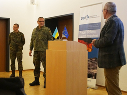 Zwei deutsche Soldaten und ein ziviler Redner bei einer sicherheitspolitischen Veranstaltung mit ukrainischen und EU-Flaggen.